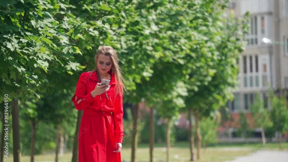 Lovely model with a cellphone. Beautiful young woman in bright red dress with a phone on the green trees background in summer. Slow motion. Lovely model with a cellphone. Beautiful young woman in bright red dress with a phone on the green trees background in summer. Slow motion.