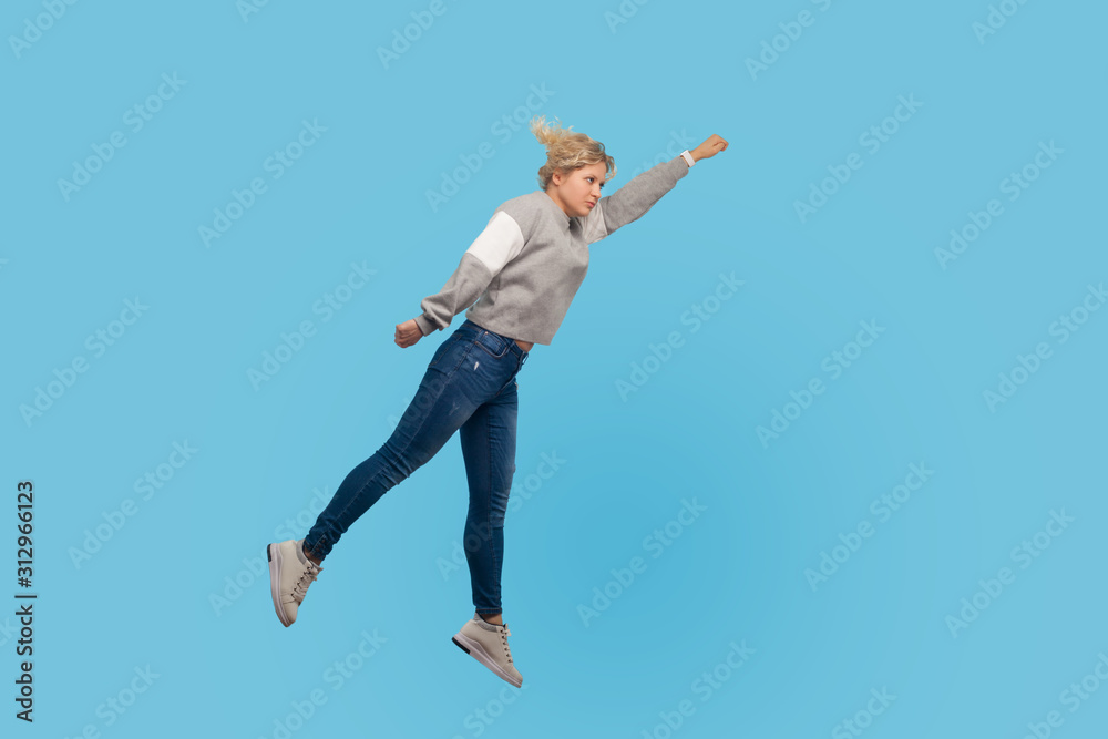 Superman. Full length portrait of determined woman in sweatshirt and jeans flying up in air, feeling superpower and freedom, trampoline to success. indoor studio shot isolated on blue background Superman. Full length portrait of determined woman in sweatshirt and jeans flying up in air, feeling superpower and freedom, trampoline to success. indoor studio shot isolated on blue background