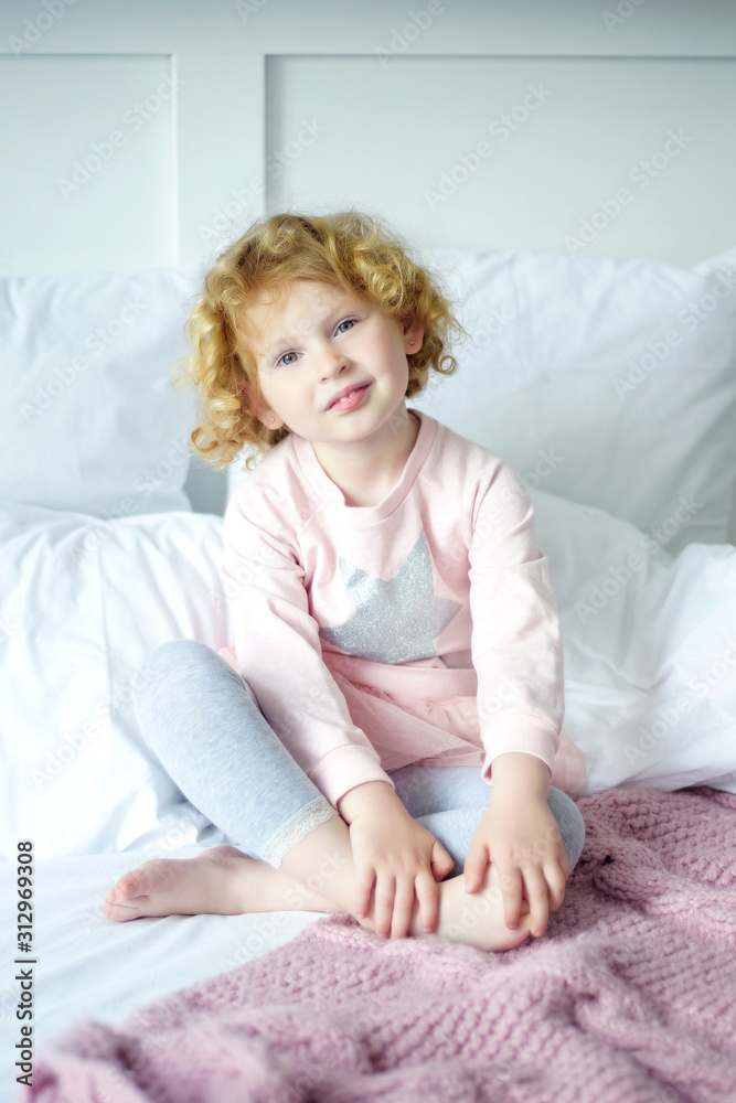 Portrair of little smiling 5 years old girl with curly blond hair sitting in pink and gray pajama in her bed. Day of sleep. Happy childhood in cozy home with parents care Portrair of little smiling 5 years old girl with curly blond hair sitting in pink and gray pajama in her bed. Day of sleep. Happy childhood in cozy home with parents care