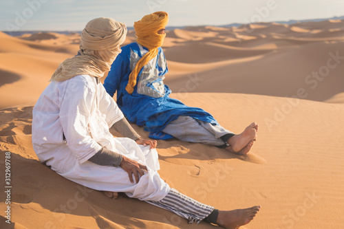 Bedouin nomad, Sahara desert, Morocco. Portrait of a Bedouin nomad with colorful turban and big smile sitting on sand dune popular tourist spot. A tuareg man portrait with his traditional clothes.