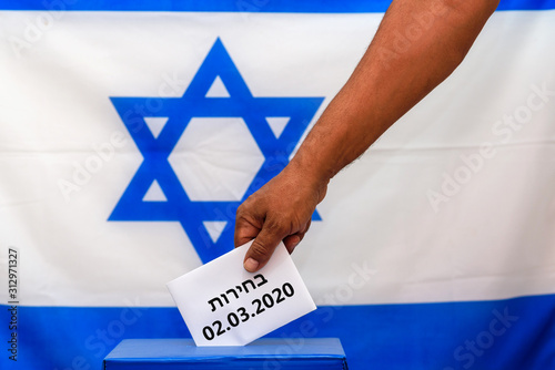 Israeli man putting a ballot paper in a ballot box on election day, third 2020 Israeli vote. Close up of hand with white votes paper on Israel flag background. Hebrew text Election 02.03.2020 photo