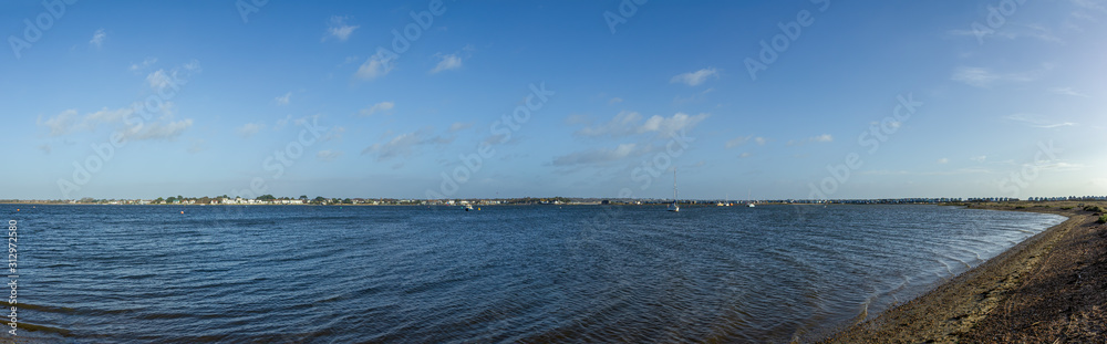A panoramic view of the Christchurch (UK) harbor with sandy beach in the foreground and boats, houses in the background under a majestic blue sky and some white clouds