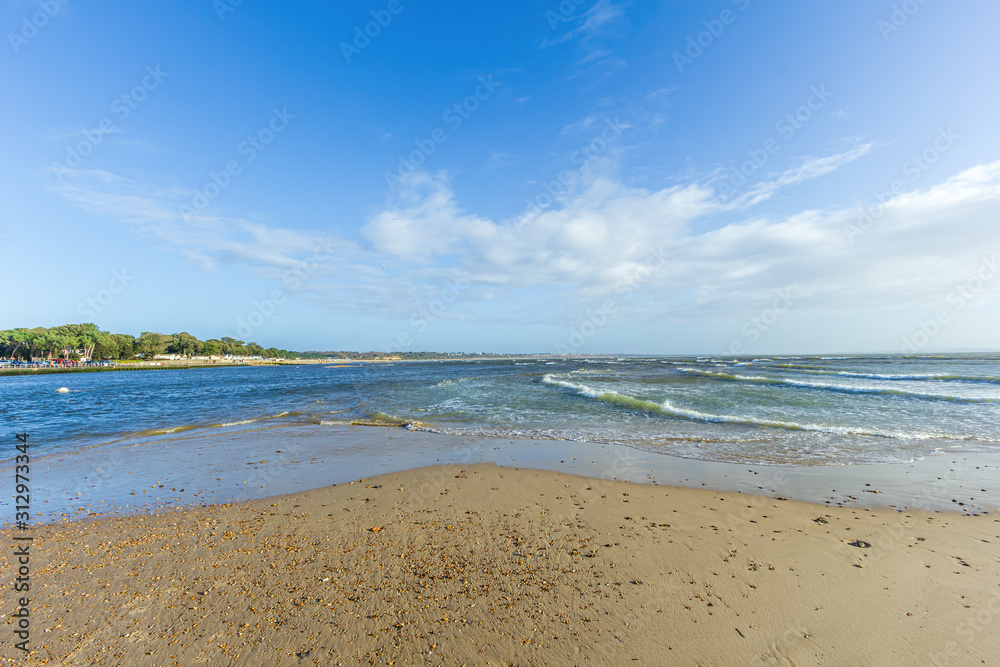 A view of a choppy water channel between Christchurch (UK) harbor and bay with sandy beach and trees along the bank under a majestic blue sky and some white clouds