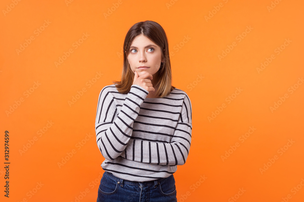 Think up plan. Portrait of pensive woman with brown hair in long sleeve striped shirt standing holding hand on chin and looking up, thinking intensely. indoor studio shot isolated on orange background Think up plan. Portrait of pensive woman with brown hair in long sleeve striped shirt standing holding hand on chin and looking up, thinking intensely. indoor studio shot isolated on orange background