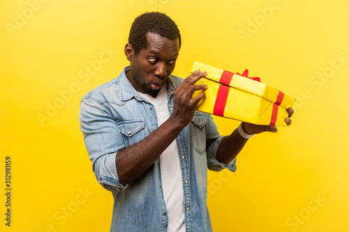 Long-awaited interesting gift! Portrait of happy man in denim casual shirt unpacking present, looking inside box with curious happy expression. indoor studio shot isolated on yellow background