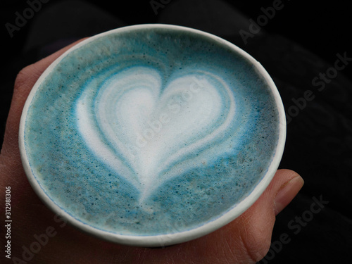 A woman hand hplding eco friendly cup of hot butterfly pea latte with beautiful heart-shaped latte art, morning refreshment