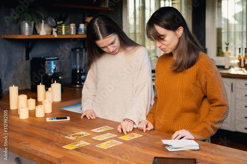 Two young femaies playing with tarot cards in a kitchen. Candles and cellphones visible