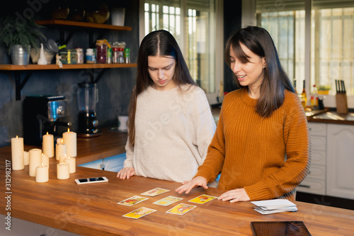 Two young femaies laying out deck of tarot cards in a kitchen. Candles and mobile devices present.
