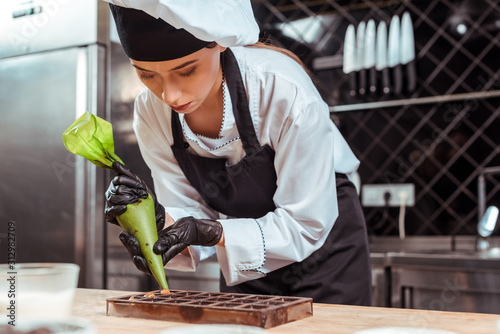 selective focus of attractive chocolatier in black latex gloves holding pastry bag near chocolate molds photo