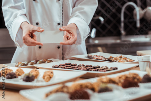 selective focus of chocolatier holding blank card in hands near tasty chocolate candies on plates