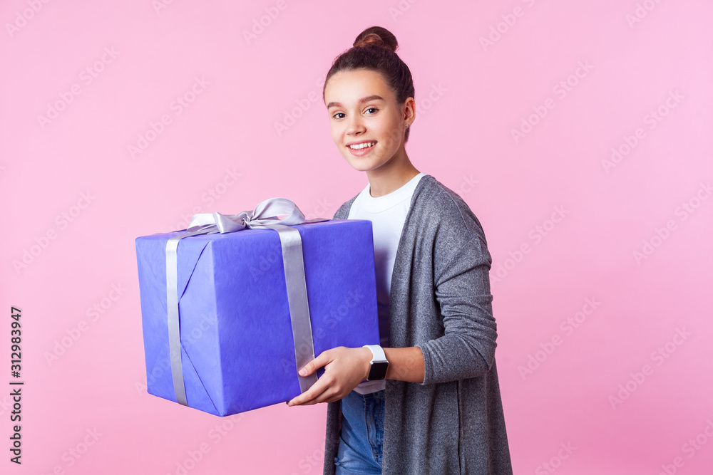 Holiday present and bonuses. Portrait of cheerful teenage brunette girl with bun hairstyle in casual clothes holding gift box and and smiling at camera. indoor studio shot isolated on pink background Holiday present and bonuses. Portrait of cheerful teenage brunette girl with bun hairstyle in casual clothes holding gift box and and smiling at camera. indoor studio shot isolated on pink background