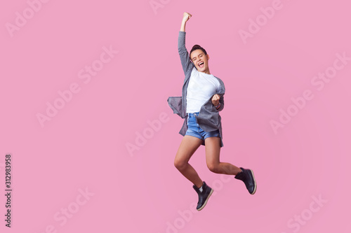 I did it! Energetic teen girl with bun hairstyle in casual clothes jumping with closed eyes and raised fists, crazy about winning, screaming and dancing with happiness in the air, celebrating success photo