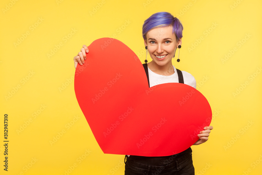 Portrait of romantic stylish hipster girl with violet short hair in overalls holding big paper heart and smiling, sharing love feeling, valentine's day. isolated on yellow background, studio shot Portrait of romantic stylish hipster girl with violet short hair in overalls holding big paper heart and smiling, sharing love feeling, valentine's day. isolated on yellow background, studio shot
