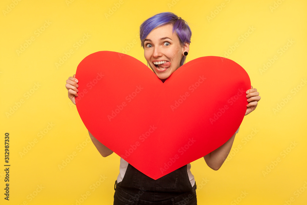 Portrait of funny cheerful hipster girl with violet short hair in overalls holding big paper heart and sticking out tongue, looking childish playful carefree. isolated on yellow background studio shot Portrait of funny cheerful hipster girl with violet short hair in overalls holding big paper heart and sticking out tongue, looking childish playful carefree. isolated on yellow background studio shot