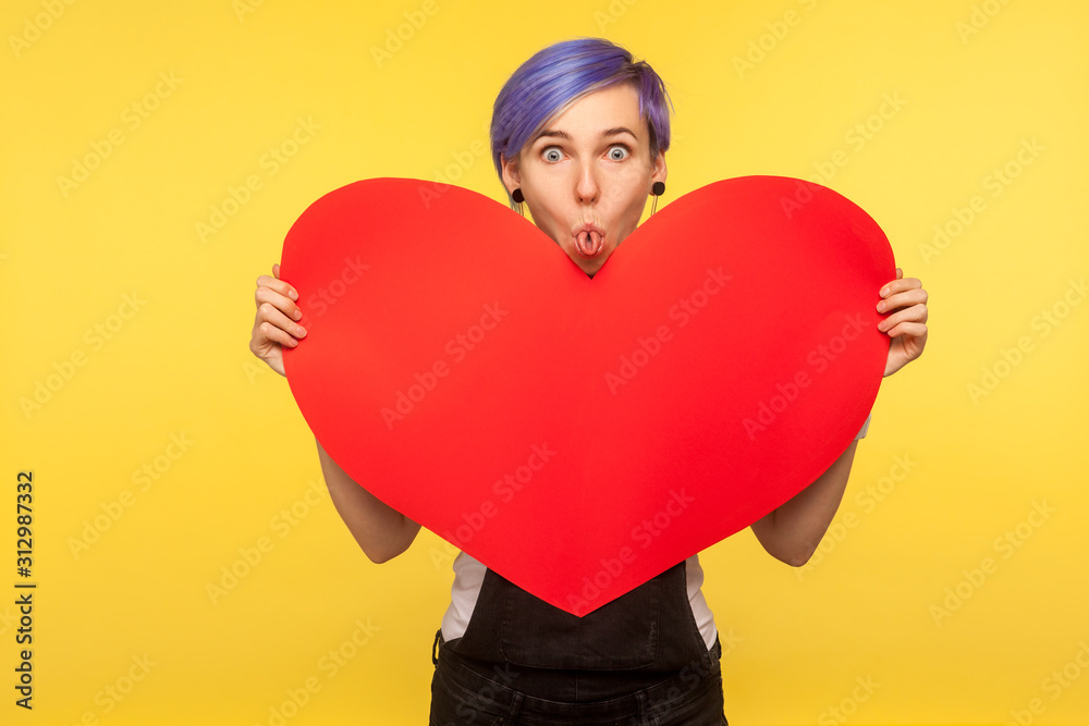 Portrait of funny amusing hipster girl with violet short hair in overalls holding big paper heart and sticking out rolled tongue, comical childish mood, making faces. yellow background studio shot Portrait of funny amusing hipster girl with violet short hair in overalls holding big paper heart and sticking out rolled tongue, comical childish mood, making faces. yellow background studio shot
