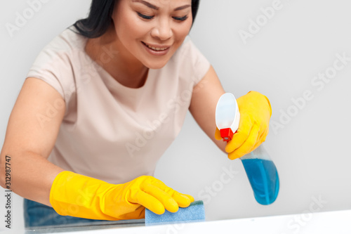 Freestyle. Asian woman in gloves standing isolated on white with spray cleaning surface with sponge smiling concentrated close-up