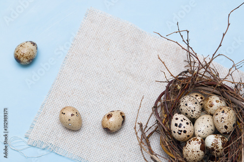 quail eggs in a nest of branches on a blue background, light fabric, Easter background, natural food, copy space, top view