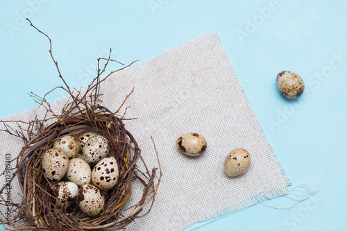 quail eggs in a nest of branches on a blue background, light fabric, Easter background, natural food, copy space, top view