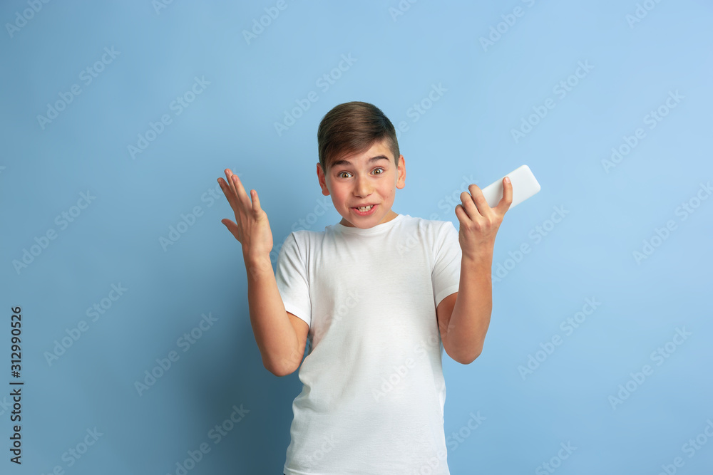 Using smartphone, looks astonished. Caucasian boy portrait isolated on blue studio background. Beautiful teen male model in white shirt posing. Concept of human emotions, facial expression, sales, ad. Using smartphone, looks astonished. Caucasian boy portrait isolated on blue studio background. Beautiful teen male model in white shirt posing. Concept of human emotions, facial expression, sales, ad.