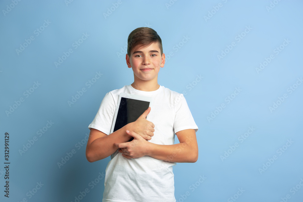 Holding tablet like student. Caucasian boy portrait isolated on blue studio background. Beautiful teen male model in white shirt posing. Concept of human emotions, facial expression, sales, ad. Holding tablet like student. Caucasian boy portrait isolated on blue studio background. Beautiful teen male model in white shirt posing. Concept of human emotions, facial expression, sales, ad.