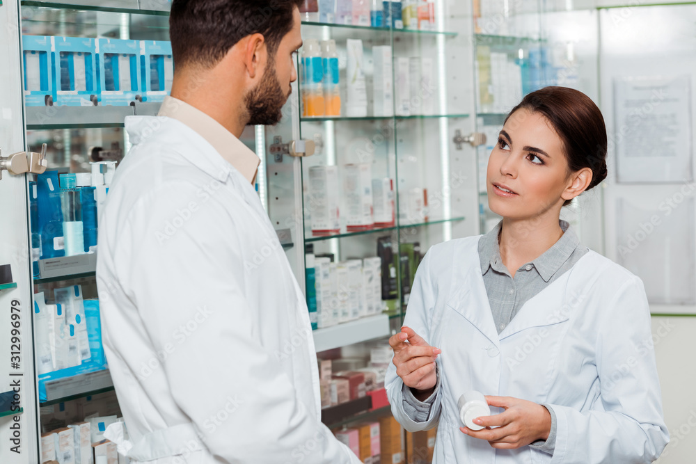 Pharmacist with jar of pills looking at colleague in pharmacy Pharmacist with jar of pills looking at colleague in pharmacy