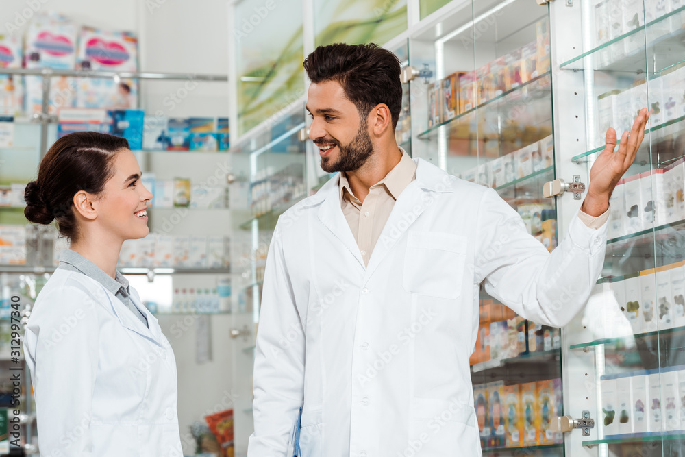 Side view of handsome pharmacist pointing at showcase to colleague in drugstore