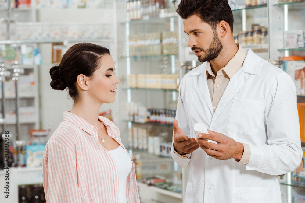 Side view of druggist showing to customer jar of pills by pharmacy showcase Side view of druggist showing to customer jar of pills by pharmacy showcase