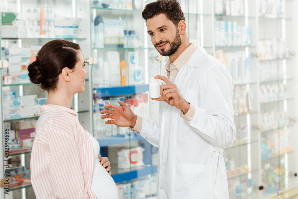 Smiling pharmacist showing to pregnant woman jar with pills by drugstore showcase