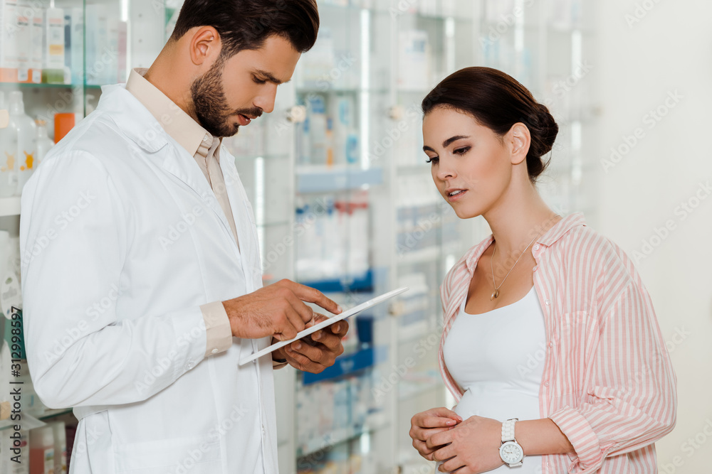 Fototapeta premium Druggist using digital tablet beside pregnant customer with medicaments on shelves at background