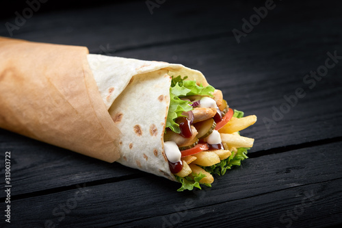 Shawarma rolled in lavash, moist grilled meat with onion, herbs and vegetables on wooden black background.
