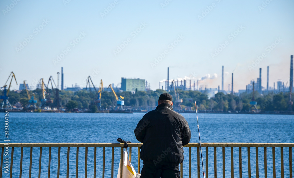 A fisherman catches fish from a bridge in the Dnipro River against the background of an industrial landscape on a fine day. Zaporizhia, Ukraine
