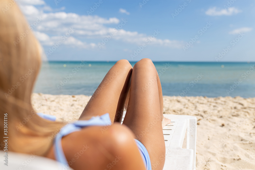The legs of a beautiful curvy blonde with long hair in a blue swimsuit gets a tan while lying in a sun lounger on a beach with a blue sea. The legs of a beautiful curvy blonde with long hair in a blue swimsuit gets a tan while lying in a sun lounger on a beach with a blue sea.