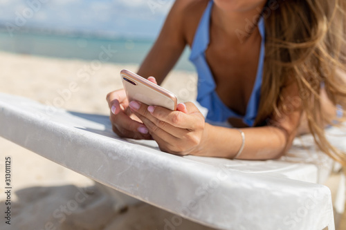 Beautiful blonde girl with long hair in a blue swimsuit uses a smartphone and sunbathes lying on a sun lounger next to the beach with a blue sea