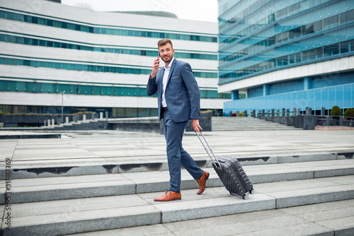 Adult blonde businessman walking with suitcase and talking on the phone