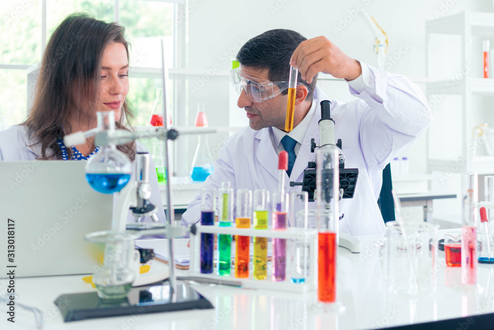 Male and Female scientist with a microscope in front of her reaching out for a test tube with blue solution in laboratory.