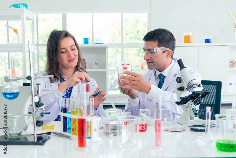 Adult caucasian woman and man chemist in protective glasses looking at microscope against chemistry lab background. Medical analysis processing concept. Adult caucasian woman and man chemist in protective glasses looking at microscope against chemistry lab background. Medical analysis processing concept.