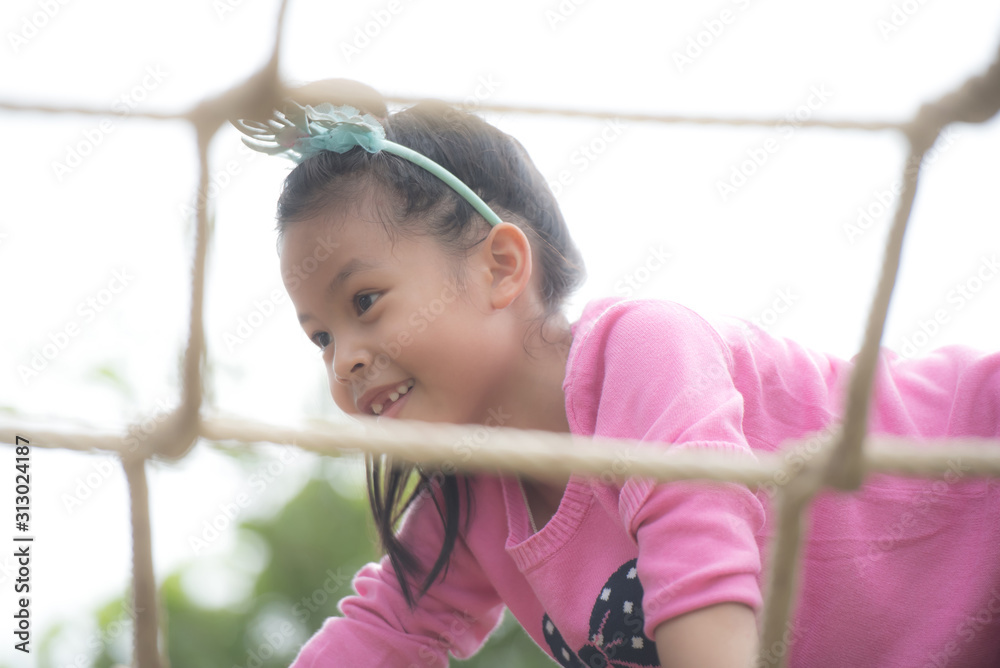 Little Girl with climbing gear climbing rope trail in an adventure park. child enjoys climbing in the ropes course adventure. Rope park. Children playing outdoors. Green tree background. Little Girl with climbing gear climbing rope trail in an adventure park. child enjoys climbing in the ropes course adventure. Rope park. Children playing outdoors. Green tree background.