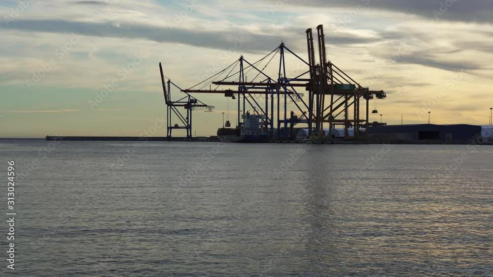 Container crane unloads cargo from a massive ship moored in the port harbor of Malaga at sunset. Costa del Sol, Spain Container crane unloads cargo from a massive ship moored in the port harbor of Malaga at sunset. Costa del Sol, Spain