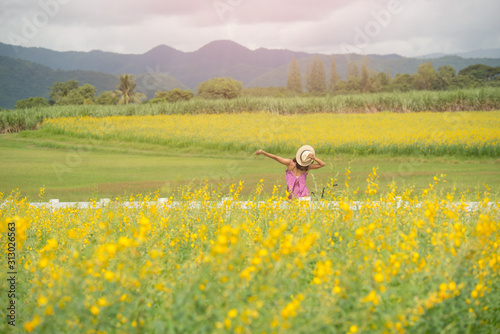 asia woman with a hat in her hand walks in a field with field flowers and smiles sincerely, happy enjoying summer in yellow field at sunset. smiling with arms raised up.  concept of freedom. © nareekarn