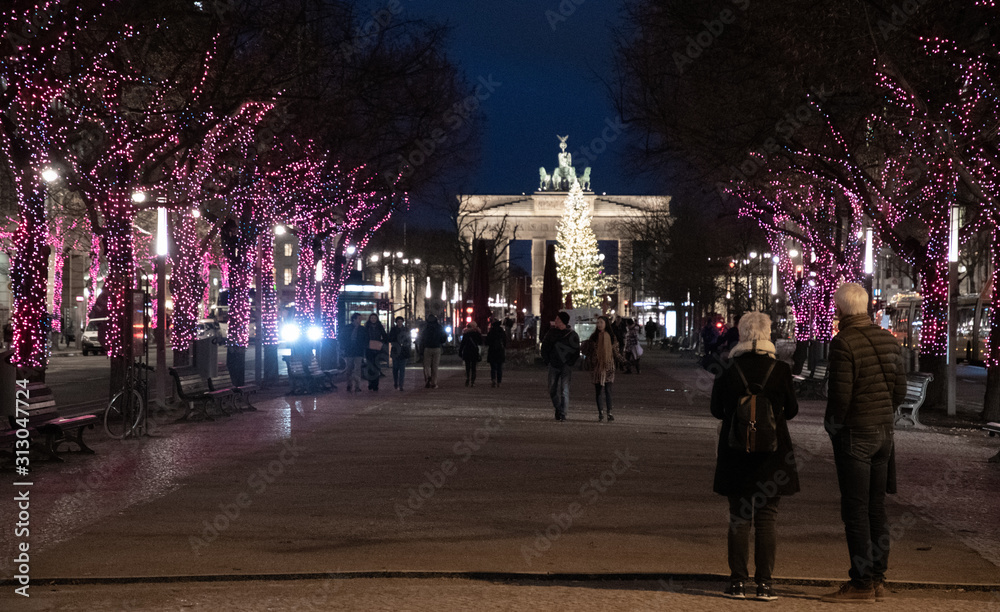 Street in city at night. Night cityscape of Unter den Linden boulevard in Berlin. Alley of illuminated trees shining in darkness and couple silhouettes. Blurred Brandenburg Gate on skyline. 