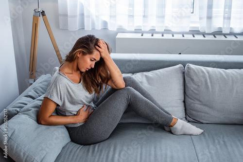 View of young woman suffering from stomachache on sofa at home. Woman sitting on bed and having stomach ache. Young woman suffering from abdominal pain while sitting on sofa at home photo