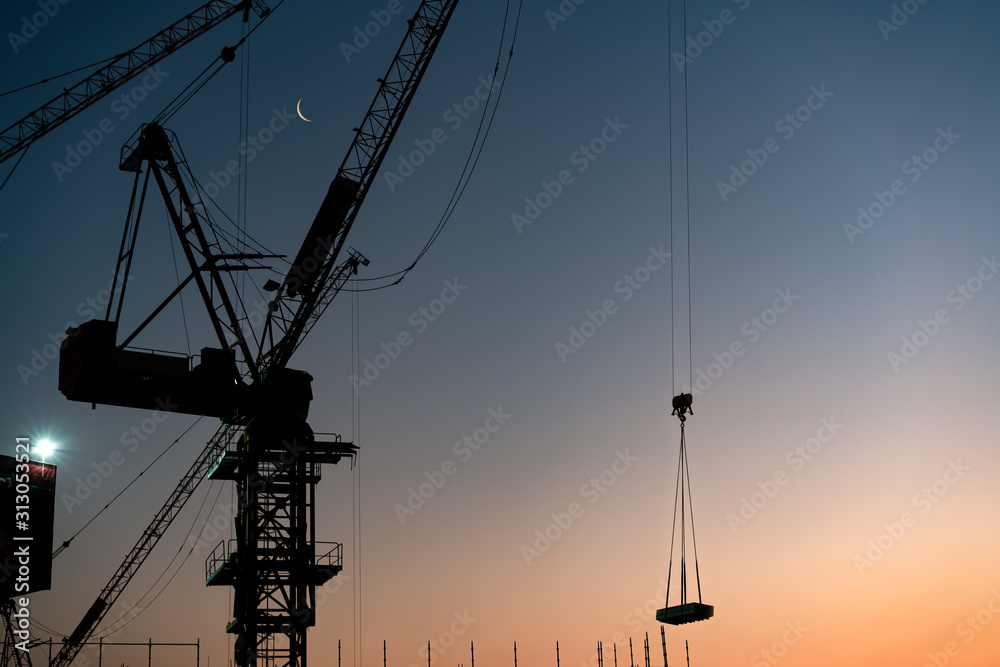 Silhouette of construction tower cranes lifting building materials during sunset - Industrial crane hoist lifting concrete at city skyscraper development - real estate, development and apartment block
