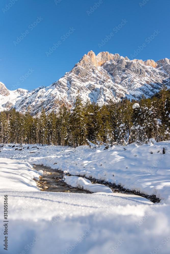 Sunny winter landscape in the alps: Mountain range, river, snowy trees, sunshine and blue sky
