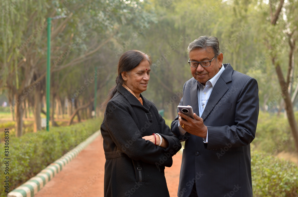 Senior Indian Bengali couple in park looking at their smart phone and smiling in a park in winters in New Delhi, India. Concept love Senior Indian Bengali couple in park looking at their smart phone and smiling in a park in winters in New Delhi, India. Concept love