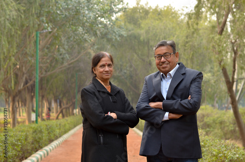 Portrait of a happy looking retired senior Indian man and woman couple smiling and posing with hands crossed / folded in a park outdoor during spring/summer season in Delhi, India. Concept love