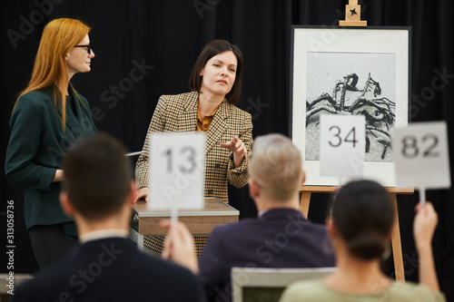 Woman standing near the painting and pointing at man who raising his sign during auction photo