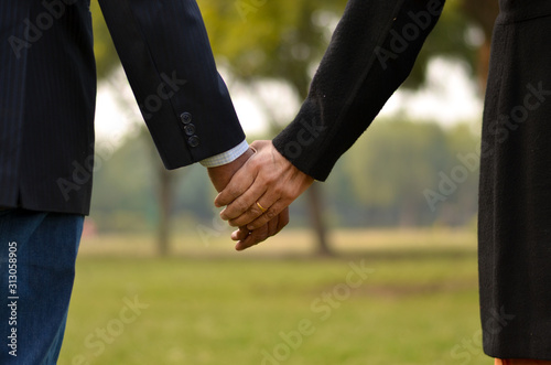 Close up cropped shot of carefree & happy elder man in black suit, holding his wife hand showing her ring who is wearing black coat in a park in winter in New Delhi, India. Concept love 
