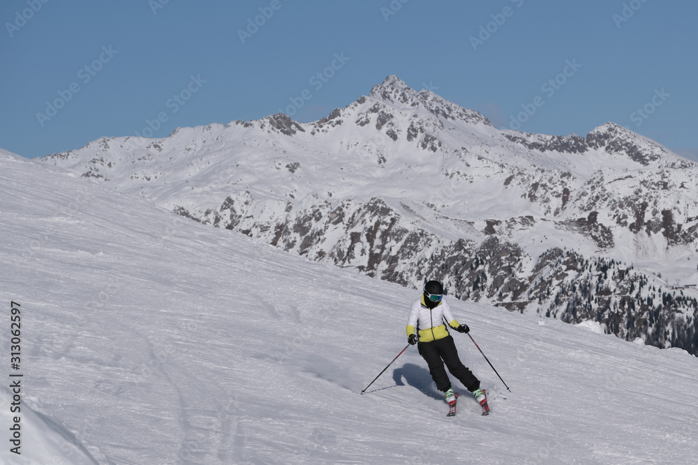 Pinzolo, Italy - December 12, 2019: Woman downhill skiing steep downhill at high speed in the background of blue sky and snowy Alps. Multicolor snowboard jacket