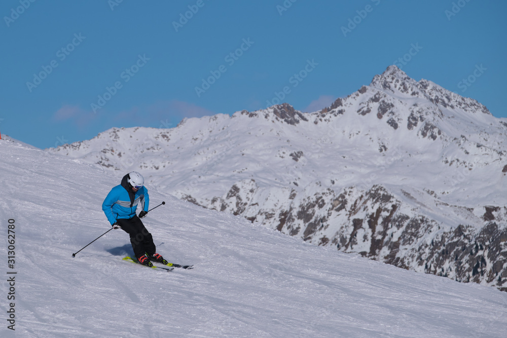 Pinzolo, Italy - December 12, 2019: Man downhill skiing steep downhill at high speed in the background of blue sky and snowy Alps. Blue ski jacket black pants