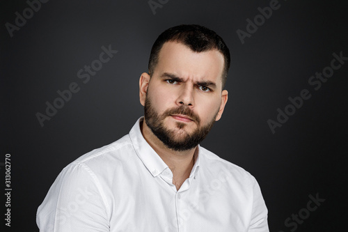man gazes steadily. Close-up portrait of handsome bearded man in white shirt looking at camera isolated on black background